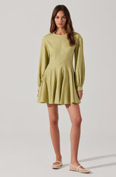 A woman is wearing a light green, long-sleeved dress with a round neckline. The dress features a fitted bodice and a flared, pleated skirt that falls above the knees. The sleeves are slightly puffed at the shoulders and gathered at the cuff.